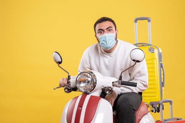 Reisconcept met verraste man met medisch masker zittend op motorfiets met gele koffer erop op geel