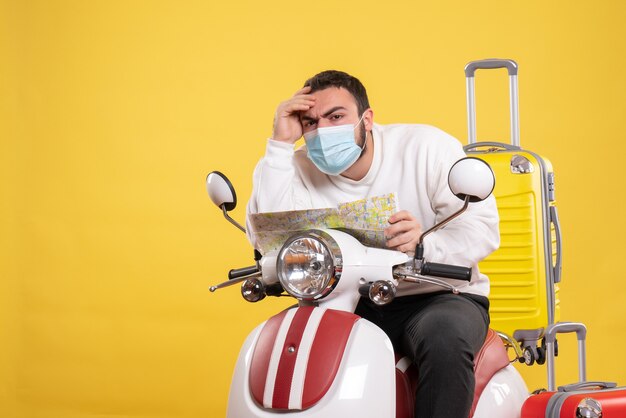 Reisconcept met verwarde jonge kerel met medisch masker zittend op motorfiets met gele koffer erop en kaart vasthoudend