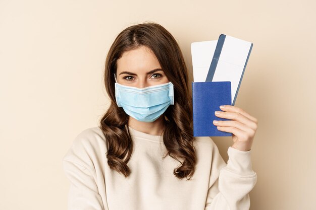 Reizen en covid-19 pandemie. Meisje met een medisch gezichtsmasker op reis, met paspoort met twee kaartjes in het vliegtuig, staande tegen een beige achtergrond.
