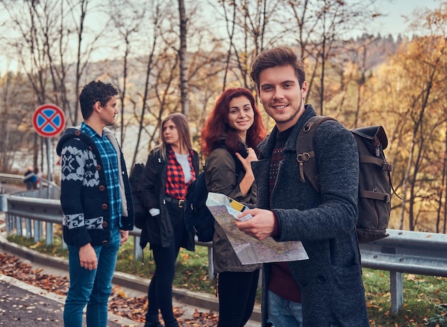 Reizen, liften, avontuur concept. groep jonge wandelaars die aan de zijlijn van de weg staan bij een prachtig herfstbos, man met kaartplanningswandeling.