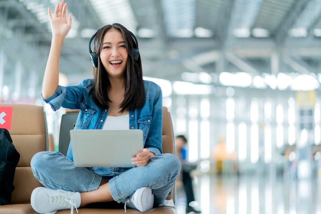 Reizen vakantie conceptpov aziatische vrouwelijke vrouw draagt hoofdtelefoon hand gebruik laptop hand golf groet naar camera met vrolijke en geluksaziatische vrouw zitten wating op wating gebied in luchthaventerminal