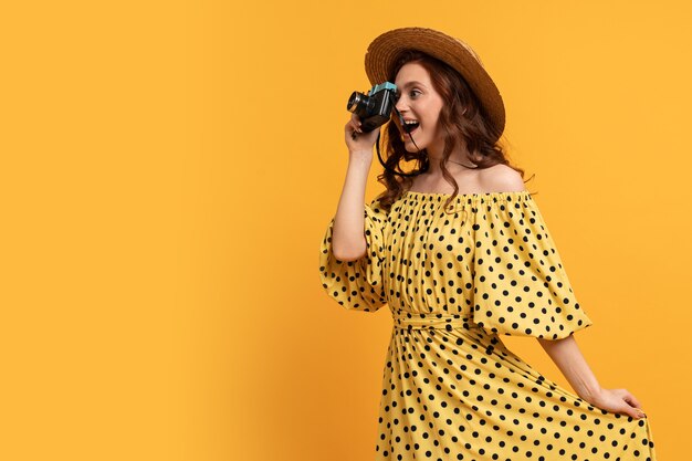 Reizende vrouw in strohoed en zomerjurk poseren met retro camera op geel