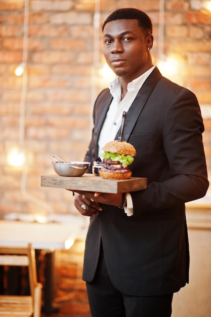 Respectabele jonge Afro-Amerikaanse man in zwart pak houdt dienblad met dubbele hamburger tegen bakstenen muur van restaurant met verlichting