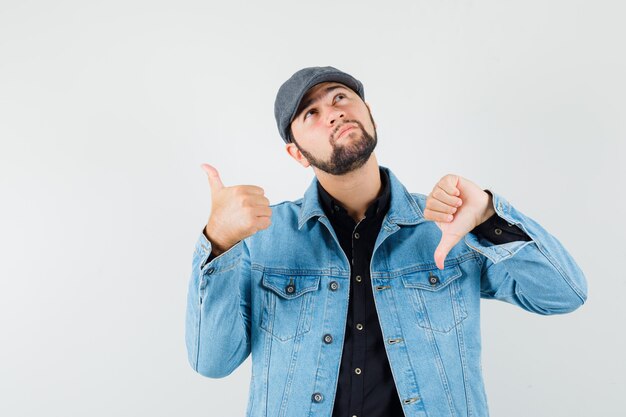 Retro-stijl man in jas, pet, shirt met duim op en neer en kijkt verward, vooraanzicht.