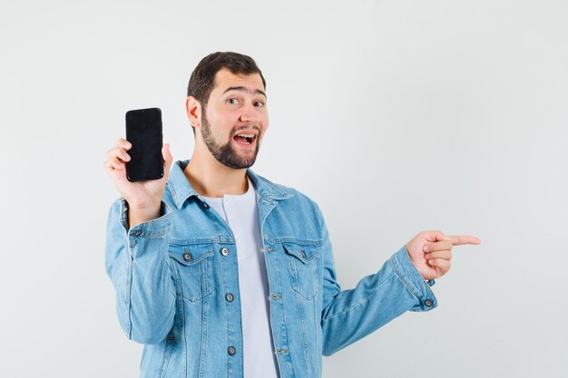 Retro-stijl man toont zijn telefoon terwijl hij opzij wijst in jasje, t-shirt en vrolijk, vooraanzicht kijkt.