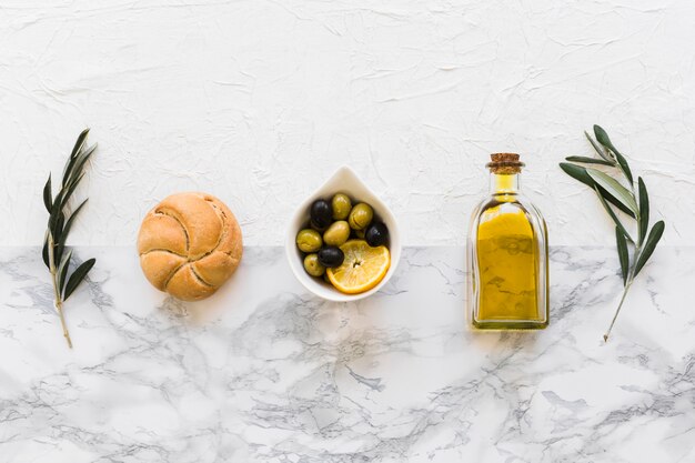 Rij van broodje, olijven en oliefles met twee takjes op wit marmer