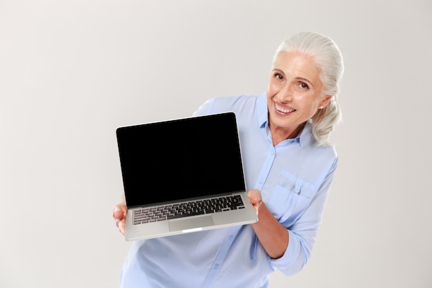 Rijpe grijs-haired vrouw die en het lege geïsoleerde scherm van laptop glimlacht toont