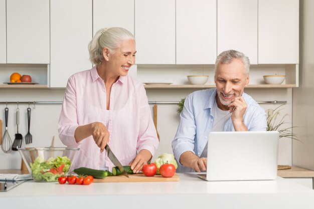 Rijpe houdende van paarfamilie gebruikend laptop en kokende salade