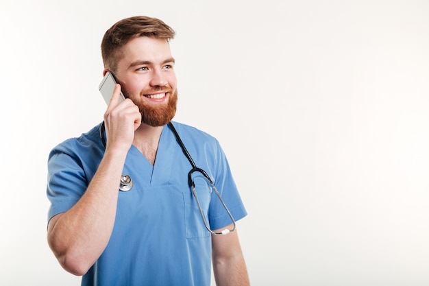 Rijpe mannelijke arts die op mobiele telefoon met glimlach spreekt terwijl status tegen witte muur
