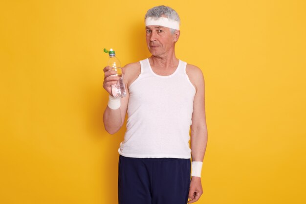Rijpe mens in hoofdband en holdingsfles water, rustend tussen sportsets, dragend t-shirt en broek