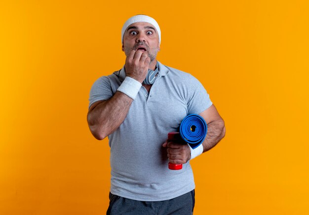 Rijpe sportieve man die in hoofdband yogamat houdt die verbaasd en verbaasd over oranje muur kijkt