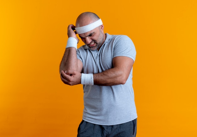 Rijpe sportieve man in hoofdband aanraken elleboog op zoek onwel gevoel pijn staande over oranje muur