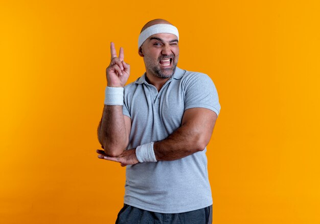 Rijpe sportieve man in hoofdband die naar de voorkant kijkt die vrolijk glimlachend overwinningsteken toont dat zich over oranje muur bevindt