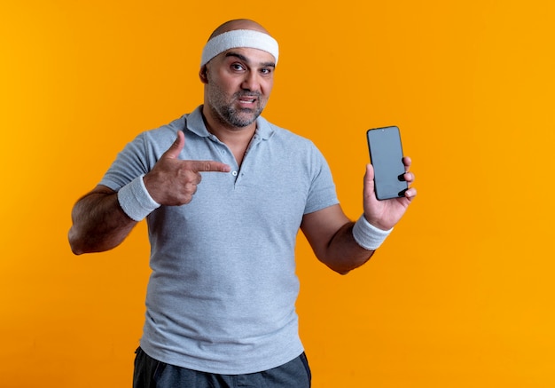 Rijpe sportieve man in hoofdband die smartphone toont die met vinger ernaar richt kijkt zelfverzekerd over oranje muur