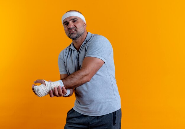 Rijpe sportieve man in hoofdband die zijn hand aanraakt die onwel kijkt met pijn die zich over oranje muur bevindt