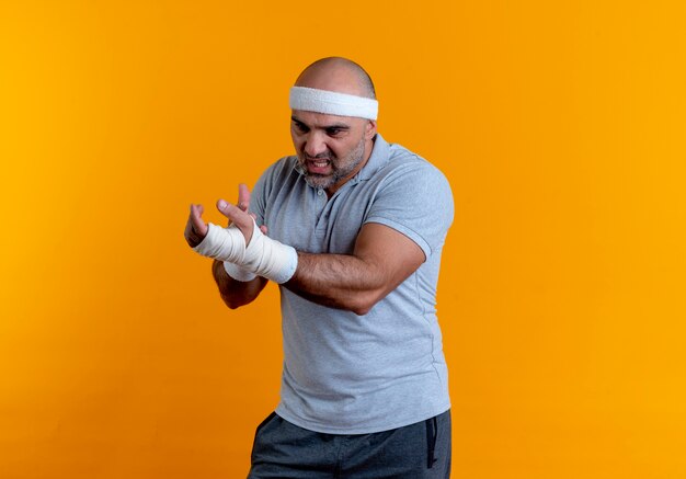 Rijpe sportieve man in hoofdband die zijn verbonden hand aanraken die onwel lijdt aan pijn die zich over oranje muur bevindt