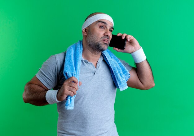 Rijpe sportieve man in hoofdband met handdoek om zijn nek op zoek verward tijdens het praten op mobiele telefoon staande over groene muur