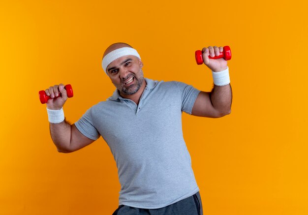 Rijpe sportieve man in hoofdband uit te werken met halters op zoek gespannen staande over oranje muur