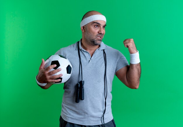 Rijpe sportieve mens in het voetbal die van de hoofdbandholding vuist opheft die opzij met ernstig gezicht kijken die zich over groene muur bevinden