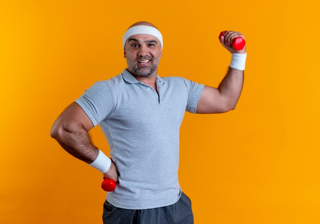 Rijpe sportieve mens in hoofdband die hand met domoor opheft die aan de voorzijde glimlachend zelfverzekerd over oranje muur kijken