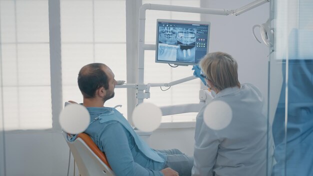 Rijpe tandarts die op tandenradiografie op monitor wijst om de dentititondiagnose uit te leggen aan patiënt met kiespijn. Tandheelkundige expert die röntgenscanresultaten toont aan man met holtepijn.