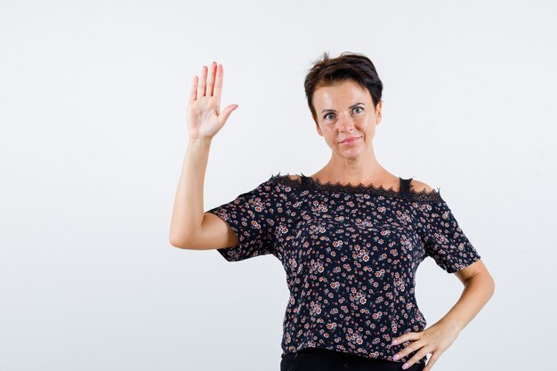 Rijpe vrouw die hand op taille houdt, stopbord in bloemenblouse, zwarte rok toont en op zoek zelfverzekerd, vooraanzicht.