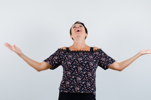 Rijpe vrouw die handen op vragende wijze in bloemenblouse en zwarte rok uitrekt en op zoek gekweld, vooraanzicht.