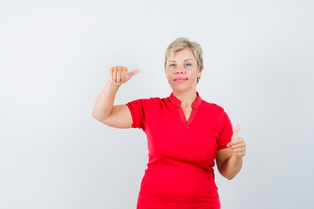 Rijpe vrouw die in rood t-shirt beweert iets vast te houden, duim opdagen.
