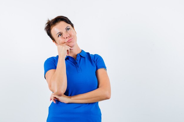 Rijpe vrouw die kin op hand in blauw t-shirt steunt en peinzend, vooraanzicht kijkt.