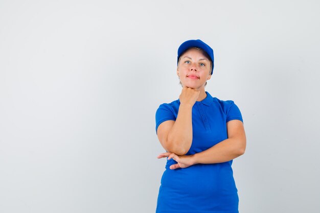 Rijpe vrouw die kin op opgeheven vuist in blauw t-shirt steunt en verstandig kijkt.