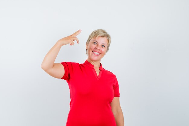 Rijpe vrouw die met hand en vingers in rood t-shirt gebaart en zelfverzekerd kijkt.