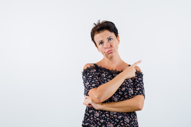 Rijpe vrouw die met wijsvinger naar rechts wijst, lippen in bloemenblouse, zwarte rok buigt en ernstig kijkt. vooraanzicht.