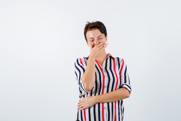 Rijpe vrouw die mond bedekt met hand, lachend, staande ogen gesloten in een gestreept overhemd en er vrolijk uitziet. vooraanzicht.