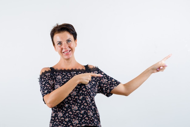 Rijpe vrouw die naar rechts wijst met wijsvingers in bloemenblouse, zwarte rok en op zoek vrolijk, vooraanzicht.