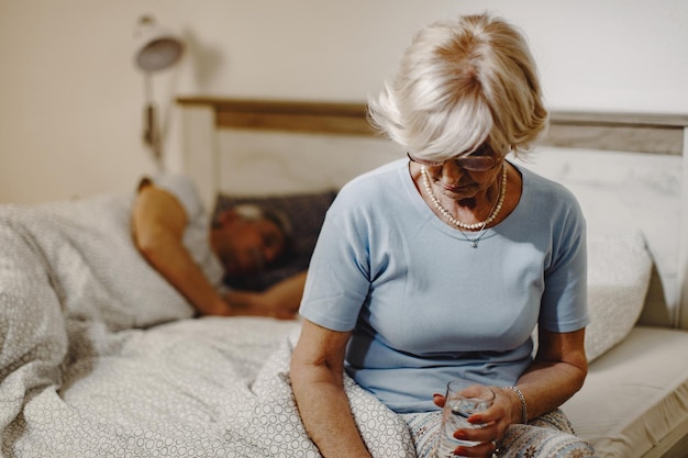 Gratis foto rijpe vrouw die op het bed zit en een glas water vasthoudt haar man slaapt op de achtergrond