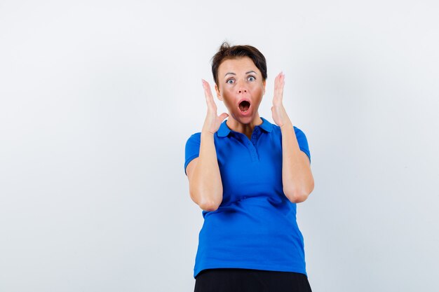 Rijpe vrouw die opgeheven handen dichtbij hoofd in blauw t-shirt houdt en geschokt kijkt. vooraanzicht.