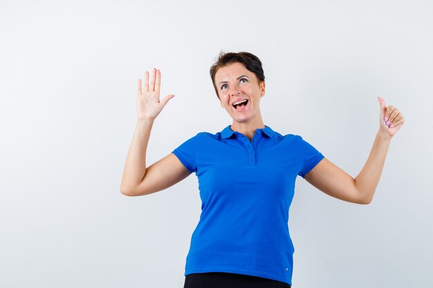 Rijpe vrouw die palm en duim in blauw t-shirt toont en gelukkig, vooraanzicht kijkt.