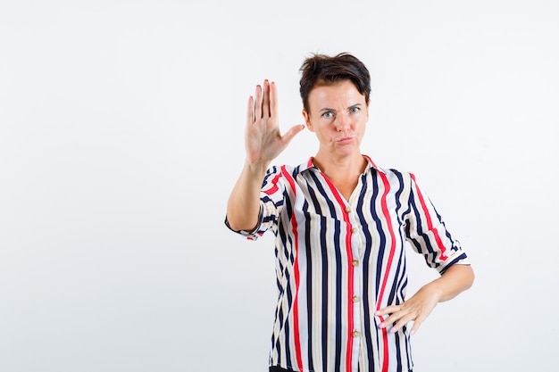 Rijpe vrouw die stopbord toont, hand op taille in gestreepte blouse houdt en ernstig, vooraanzicht kijkt.