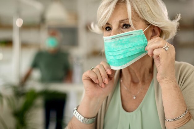 Rijpe vrouw die thuis beschermend gezichtsmasker gebruikt tijdens coronavirusepidemie