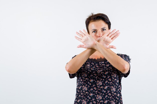 Rijpe vrouw die twee gekruiste armen houdt, geen teken in bloemenblouse, zwarte rok gebaart en ernstig kijkt. vooraanzicht.