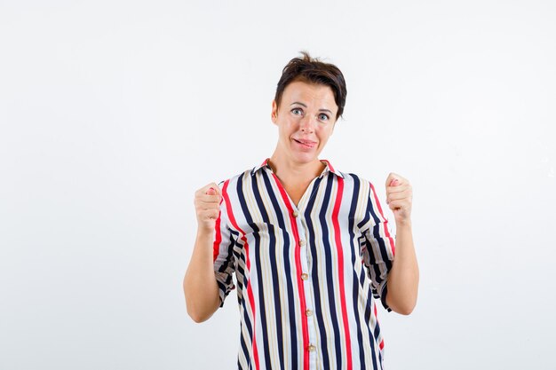 Rijpe vrouw die vijgengebaar toont, tong uitsteekt in gestreepte blouse en geamuseerd kijkt. vooraanzicht.