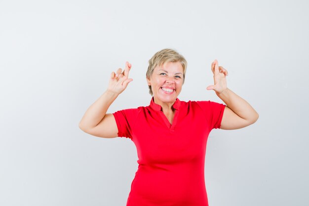 Rijpe vrouw die vingers gekruist in rood t-shirt houdt en vrolijk kijkt