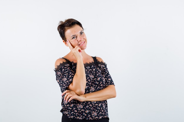 Rijpe vrouw die wang op palm leunt, over iets in bloemenblouse, zwarte rok denkt en gelukkig kijkt. vooraanzicht.