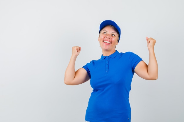 Rijpe vrouw die winnaargebaar in blauw t-shirt toont en gelukkig kijkt.