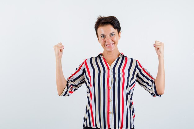 Rijpe vrouw die winnaargebaar in gestreepte blouse toont en drijvend kijkt. vooraanzicht.