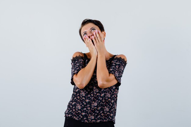 Rijpe vrouw hand in hand op de wangen tijdens het kijken in blouse en op zoek verbaasd, vooraanzicht.