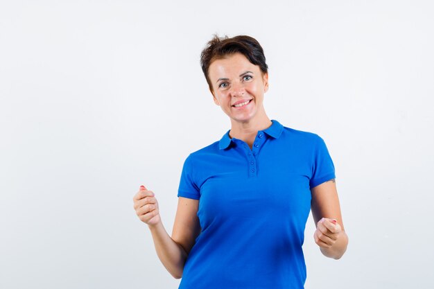 Rijpe vrouw in blauw t-shirt die winnaargebaar toont en blij, vooraanzicht kijkt.