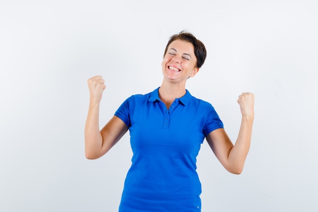 Rijpe vrouw in blauw t-shirt die winnaargebaar toont en gelukkig, vooraanzicht kijkt.