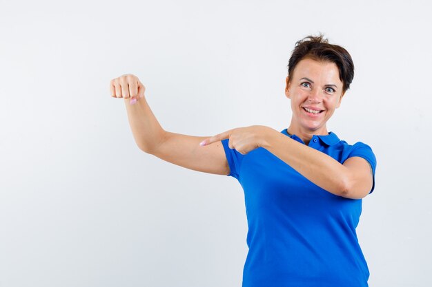 Rijpe vrouw in blauw t-shirt wijzend op de spieren van de arm en op zoek trots, vooraanzicht.