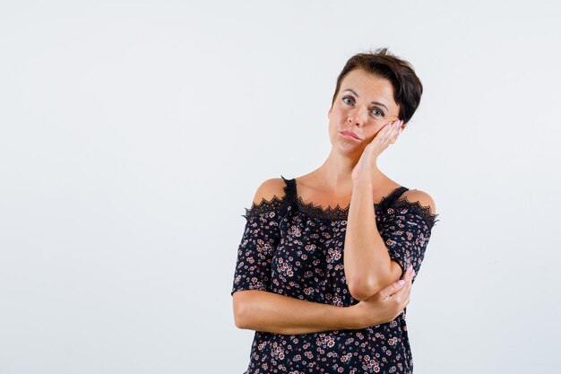 Rijpe vrouw in bloemenblouse en zwarte rok die wang op handpalm leunen, aan iets denken en peinzend, vooraanzicht kijken.